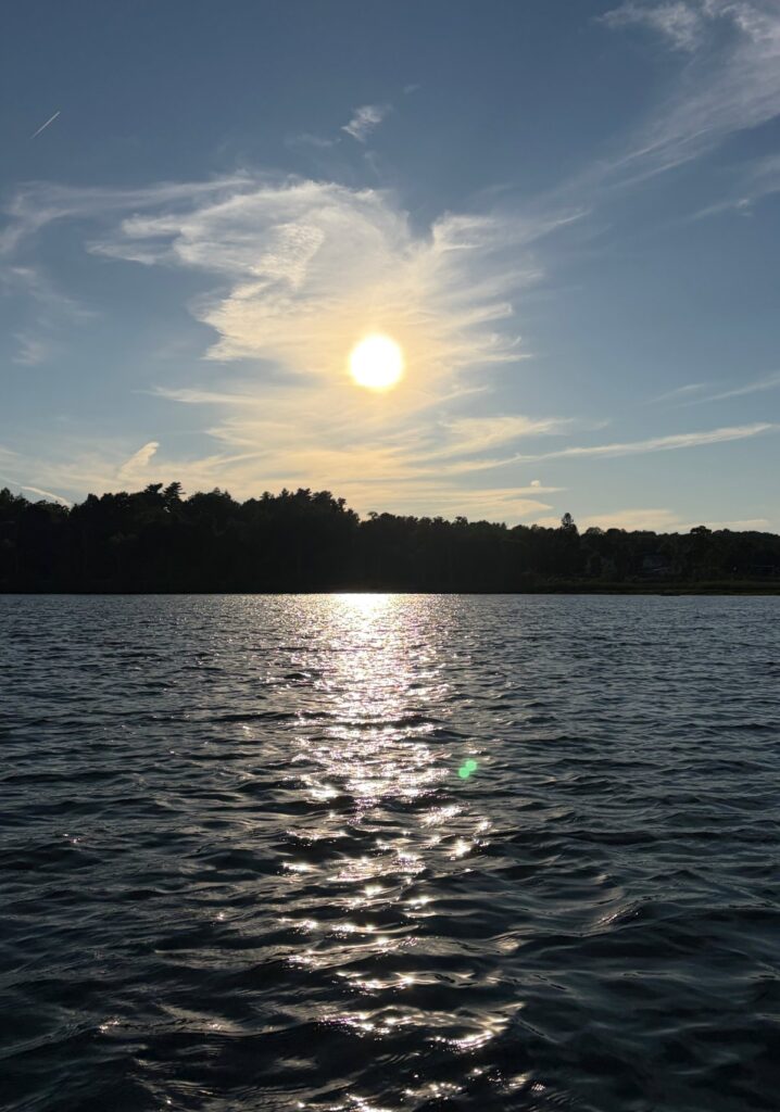 Bright sun shining over Round Lake with shimmering silver reflection on dark water, treeline in background