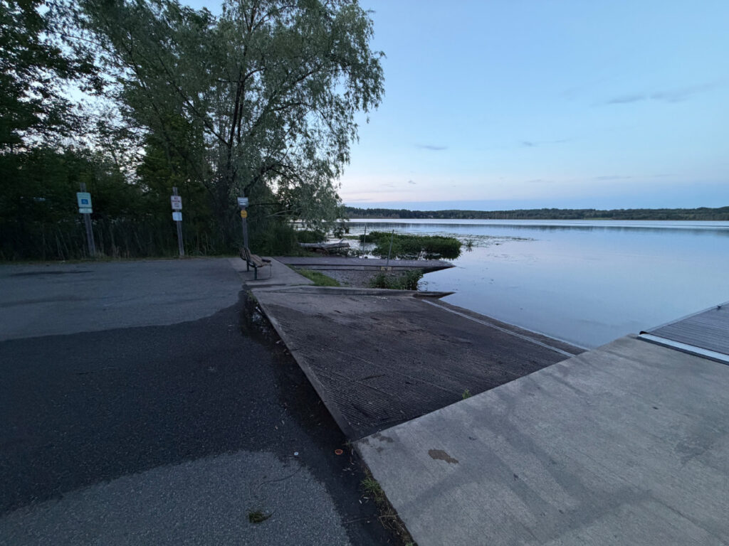 Concrete boat ramp descending into calm Round Lake at dusk, parking area with accessible parking sign visible to left, willow tree framing scene