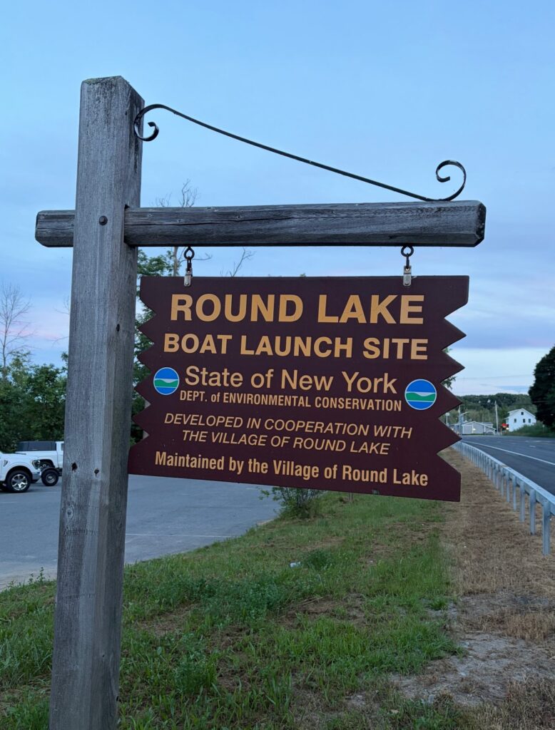 Brown wooden sign with yellow lettering reading 'ROUND LAKE BOAT LAUNCH SITE' mounted on weathered post, State of New York Department of Environmental Conservation logos visible.