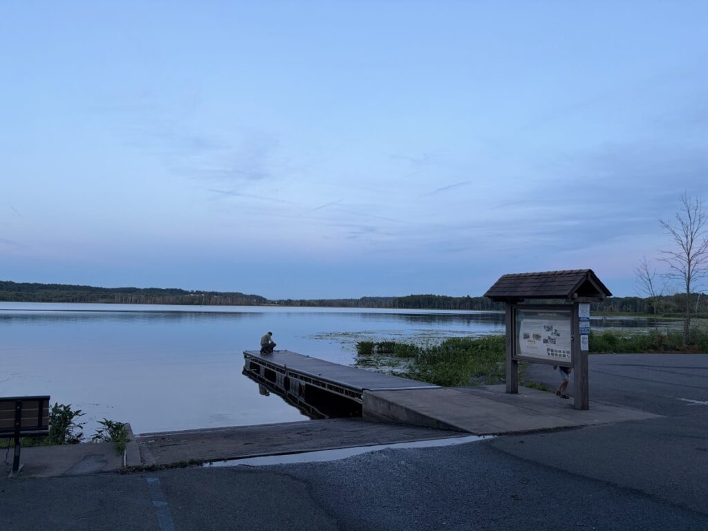 Floating dock extending into Round Lake at dusk, person sitting at dock edge, wooden information kiosk with covered roof on right