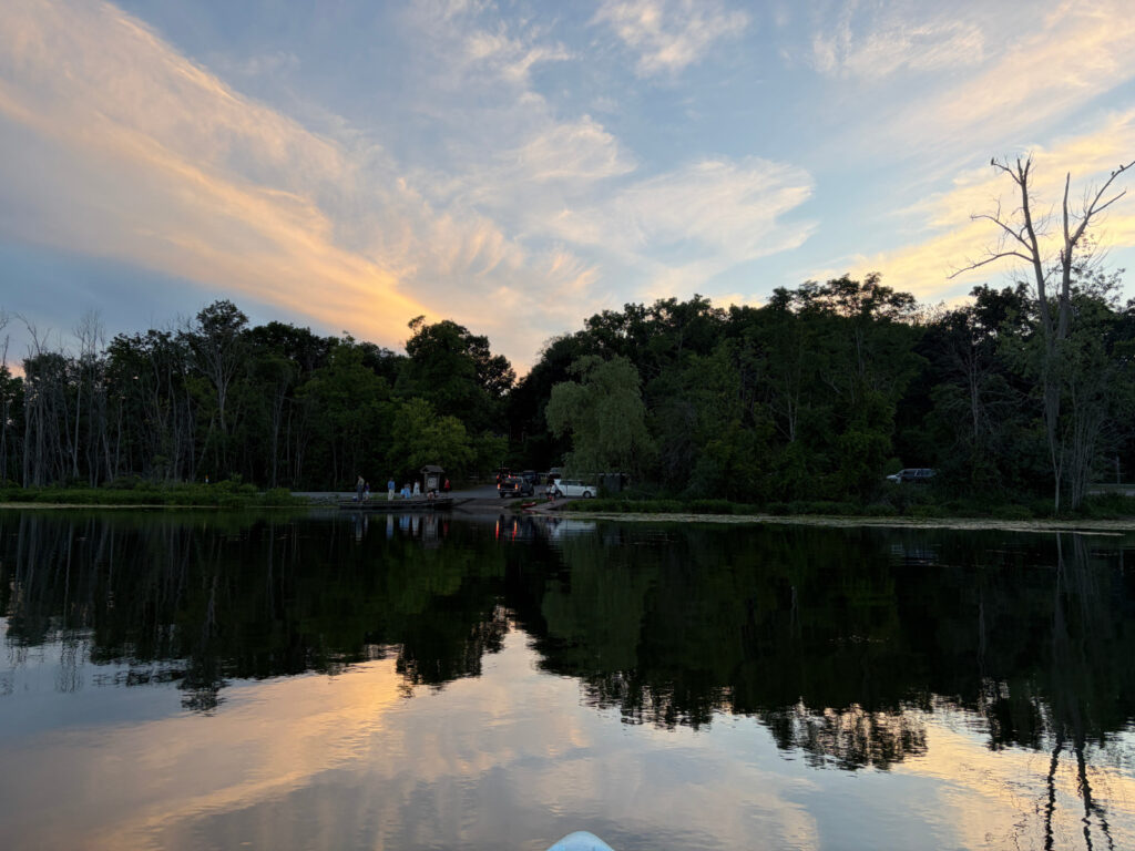 View from paddleboard looking toward Round Lake boat launch at sunset, calm water reflecting the wispy clouds against the blue sky, vehicles and people visible at dock