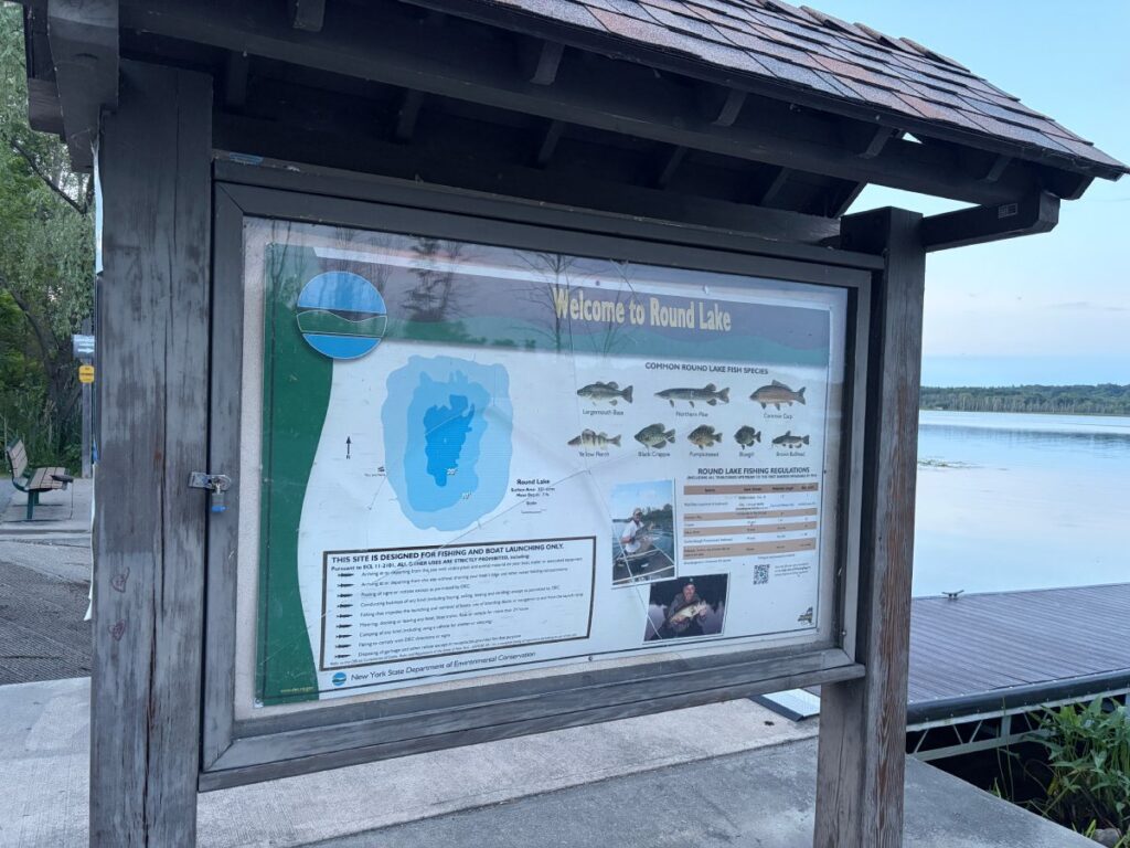 Wooden information kiosk displaying 'Welcome to Round Lake' poster with lake depth map, six illustrated fish species, and printed fishing regulations table