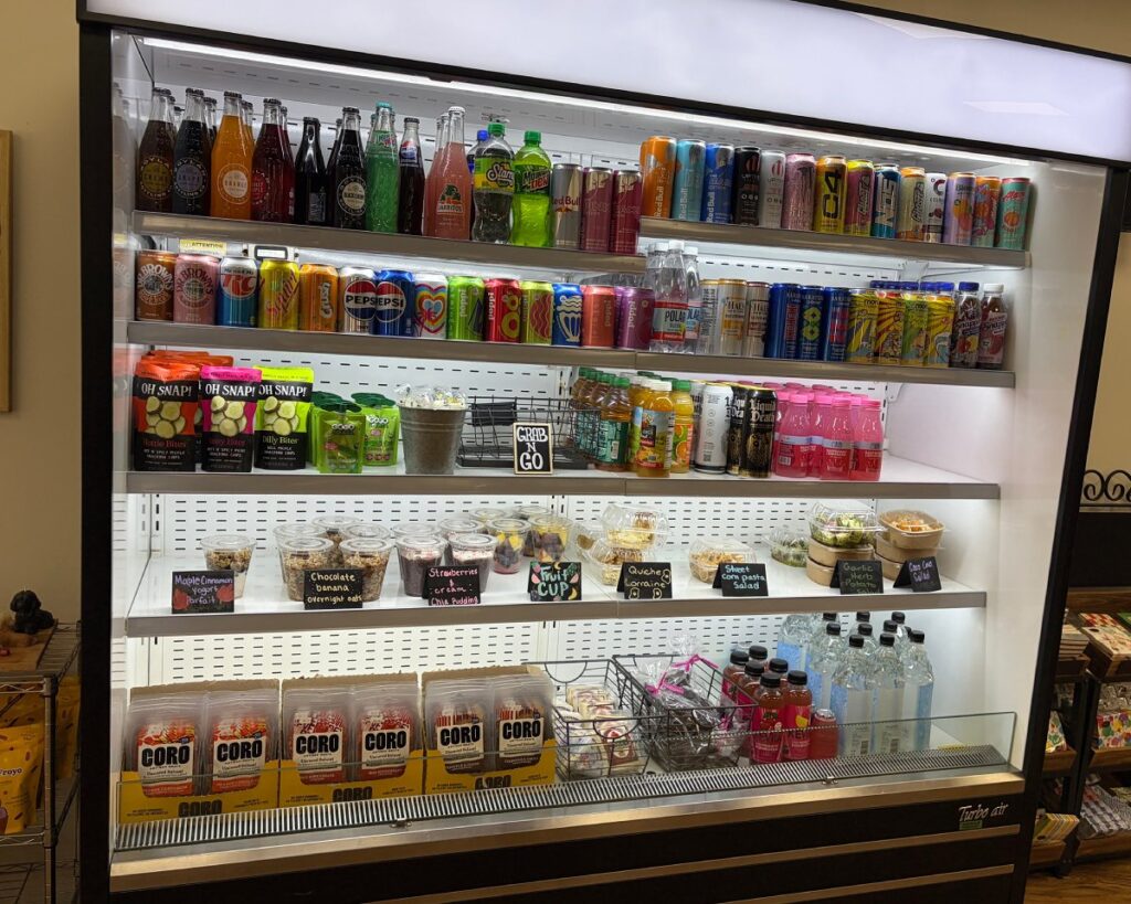 Refrigerated display cooler stocked with grab-and-go items including sodas, energy drinks, yogurt parfaits, overnight oats, salads, chips, and packaged snacks with labeled price signs.