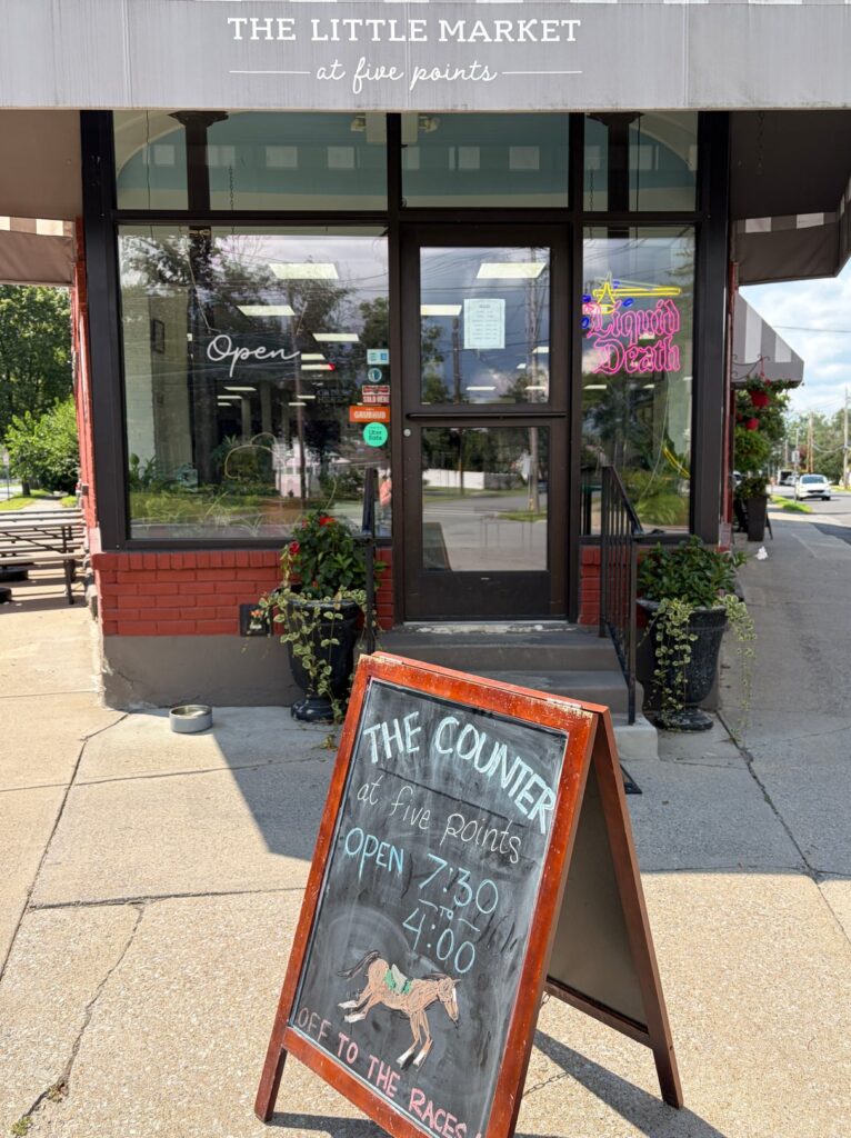 Exterior of The Counter at Five Points in Saratoga Springs, featuring a chalkboard A-frame sign displaying business hours and storefront with brick façade and potted plants flanking the entrance.