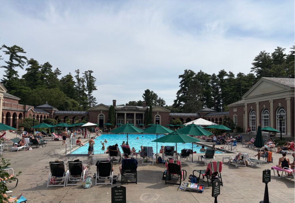 Swimming pool surrounded by chaise lounge chairs, dark green umbrellas, trees and brick arches.