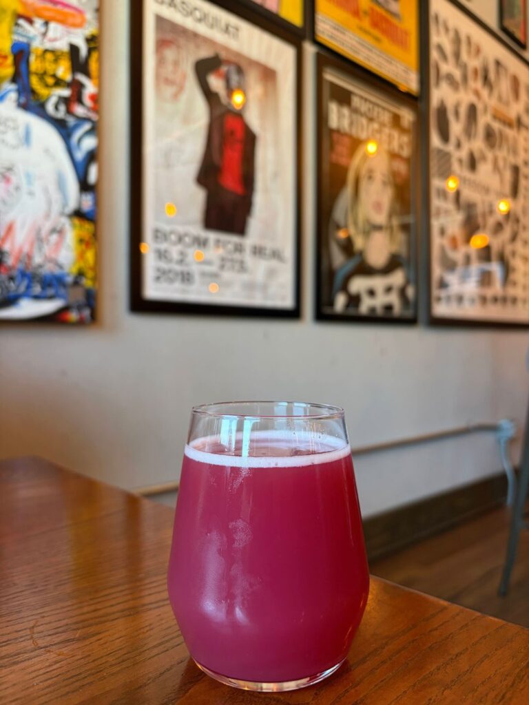A pink sour beer on a wooden table in front of a wall of framed artwork at Whitman Brewing in Saratoga.